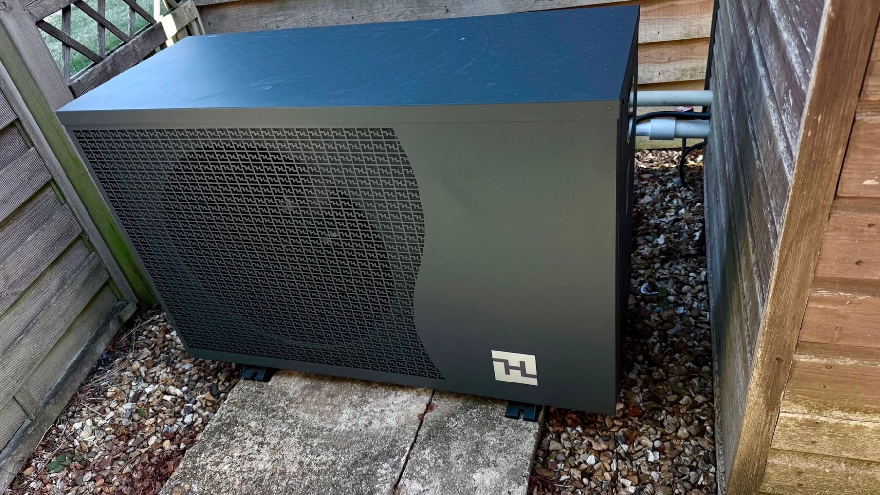 Swimming pool heat pump installed on a paved base beside a garden fence